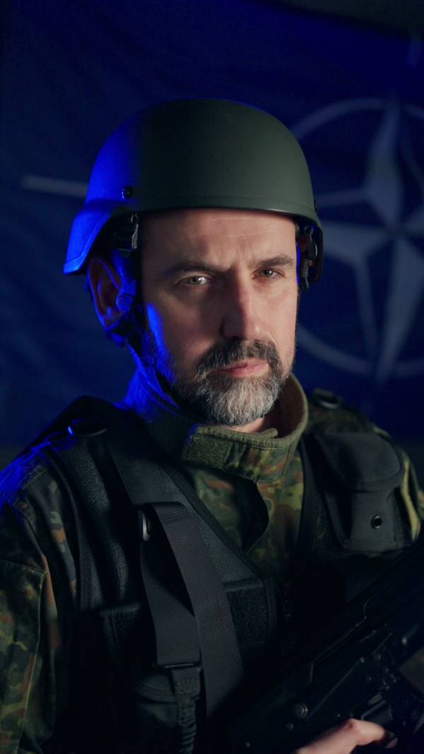 A soldier in military uniform and helmet holding weapon with NATO flag in background.