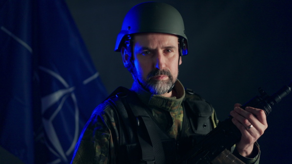 A soldier in military uniform and helmet holding weapon with NATO flag in background.