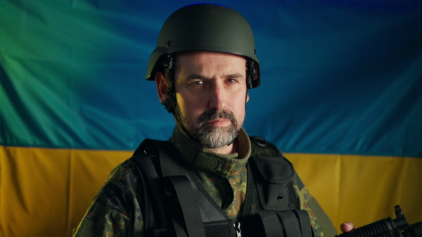 A close-up of Ukrainian soldier in military uniform and helmet looking at camera with flag at background.