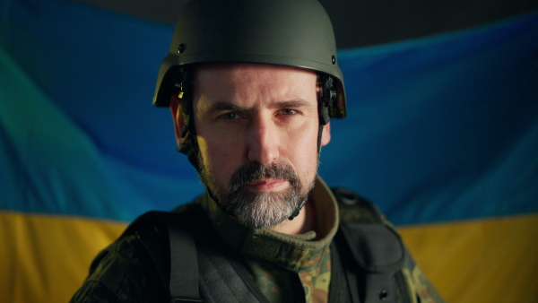 A close-up of Ukrainian soldier in military uniform and helmet looking at camera with flag at background.