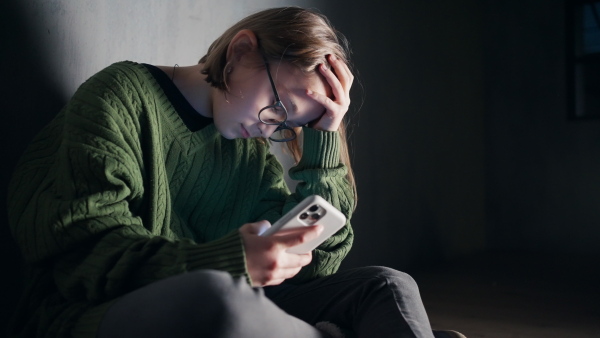 A sad little girl, alone in darkness, sitting and using smartphone, fake news and disinformation concept.