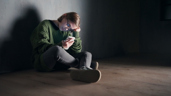 A sad little girl, alone in darkness, sitting and using smartphone, fake news and disinformation concept.