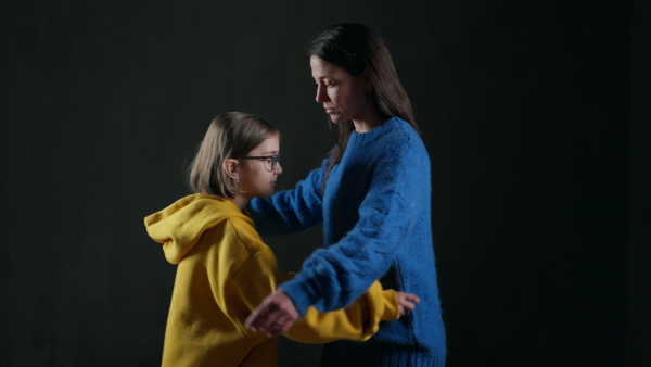 A sad refugee mother hugging her daughter, both wearing Ukrainian national colors on black background.