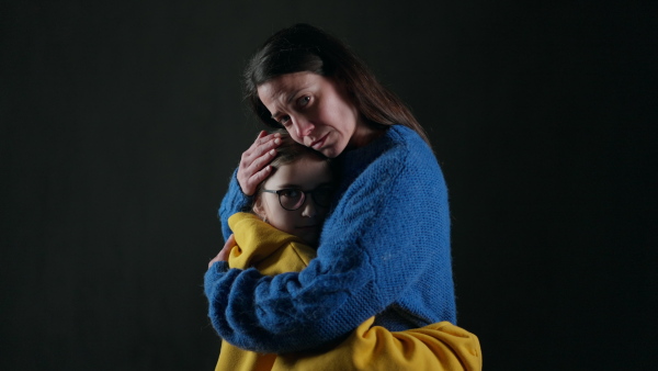 A sad refugee mother hugging her daughter, both wearing Ukrainian national colors on black background.