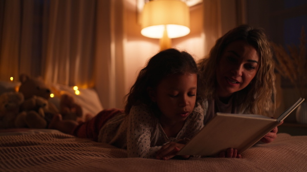 A happy mother with her little daughter lying on bed and reading book in evening at home.