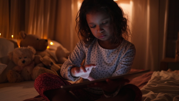 A happy little multiracial girl using tablet on bed in evening at home.