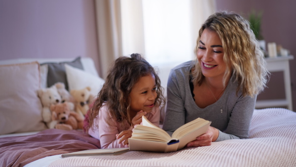 A happy mother with her little daughter lying on bed and reading book at home.