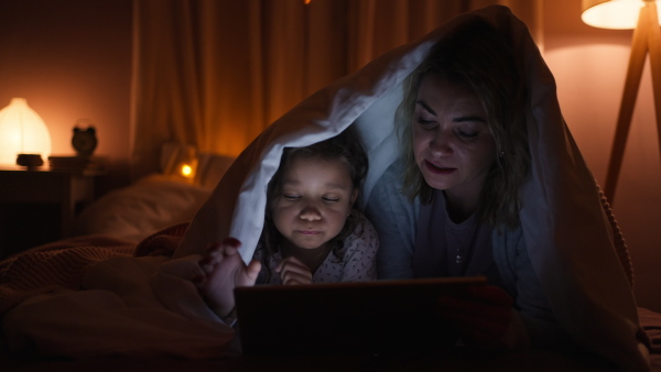 A happy mother with her little daughter lying under blanket and watching movie on tablet at home.