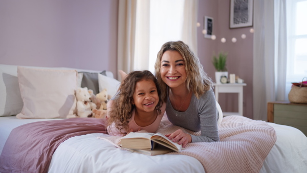 A happy mother with her little daughter lying on bed and reading book at home.