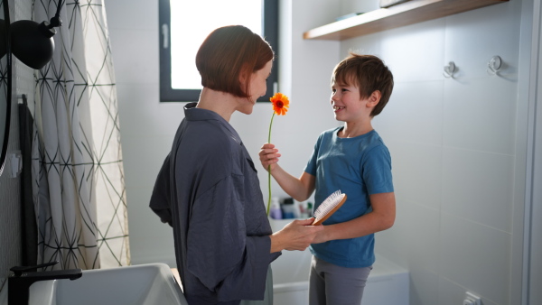 A lttle boy congratulates mother and gives her flower in bathroom at home.