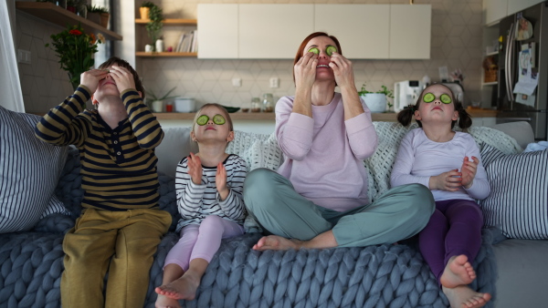 Three little children with a mother sitting on sofa and putting cucumber on faces at home.