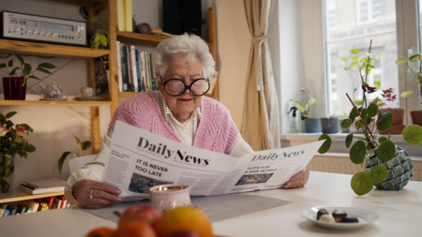 A senior woman with funny glasses reading newspaper at home and looking at camera