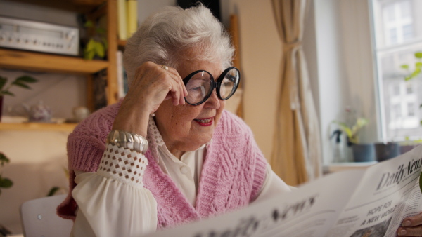 A senior woman with funny glasses reading newspaper at home and looking at camera