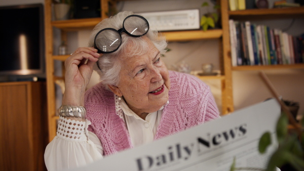 A senior woman with funny glasses reading newspaper at home and looking at camera