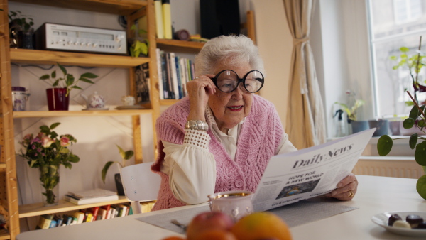 A senior woman with funny glasses reading newspaper at home and looking at camera