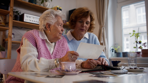 A senior mother with her adult daughter looking at old family pictures at home together