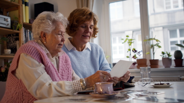 A senior mother with her adult daughter looking at old family pictures at home together