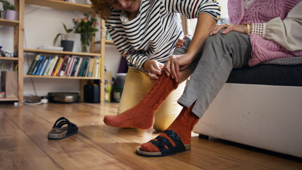 A nursing caregiver helps put on slippers to senior woman at home.