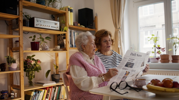 A senior mother with her adult daughter reading newspaper at home together