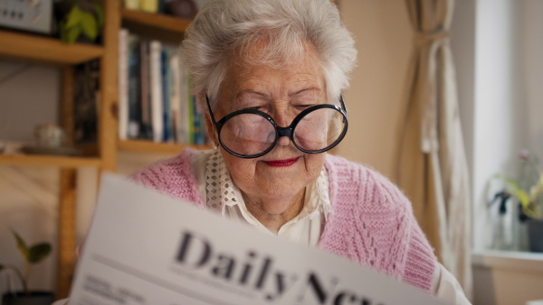 A senior woman with funny glasses reading newspaper at home and looking at camera