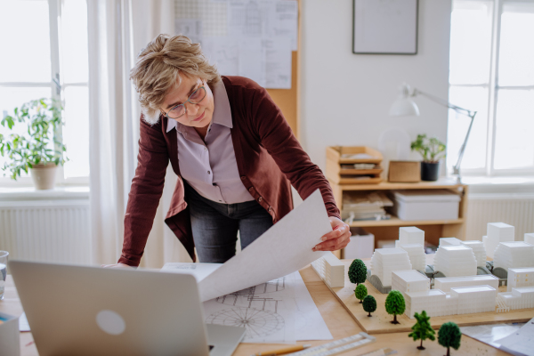 A senior woman architect with model of houses looking at blueprints in office.