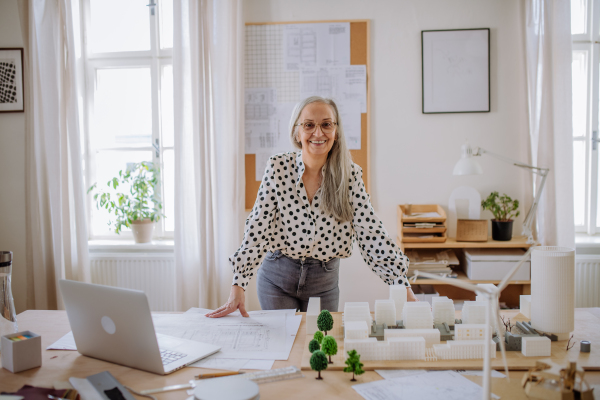 Happy senior woman architect with model of houses standing in office and looking at camera.