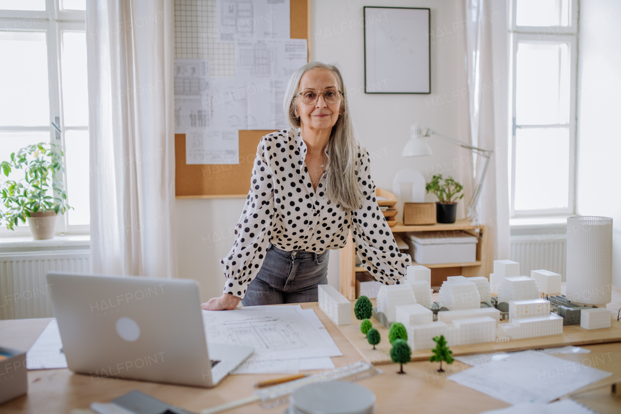 Happy senior woman architect with model of houses standing in office and looking at camera.