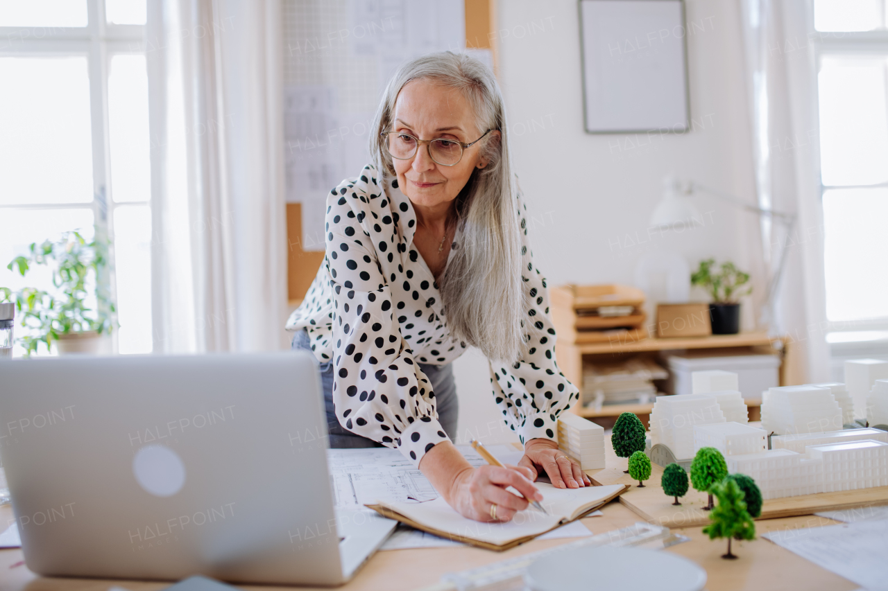 A senior woman architect with model of houses standing in office and and writing notes.