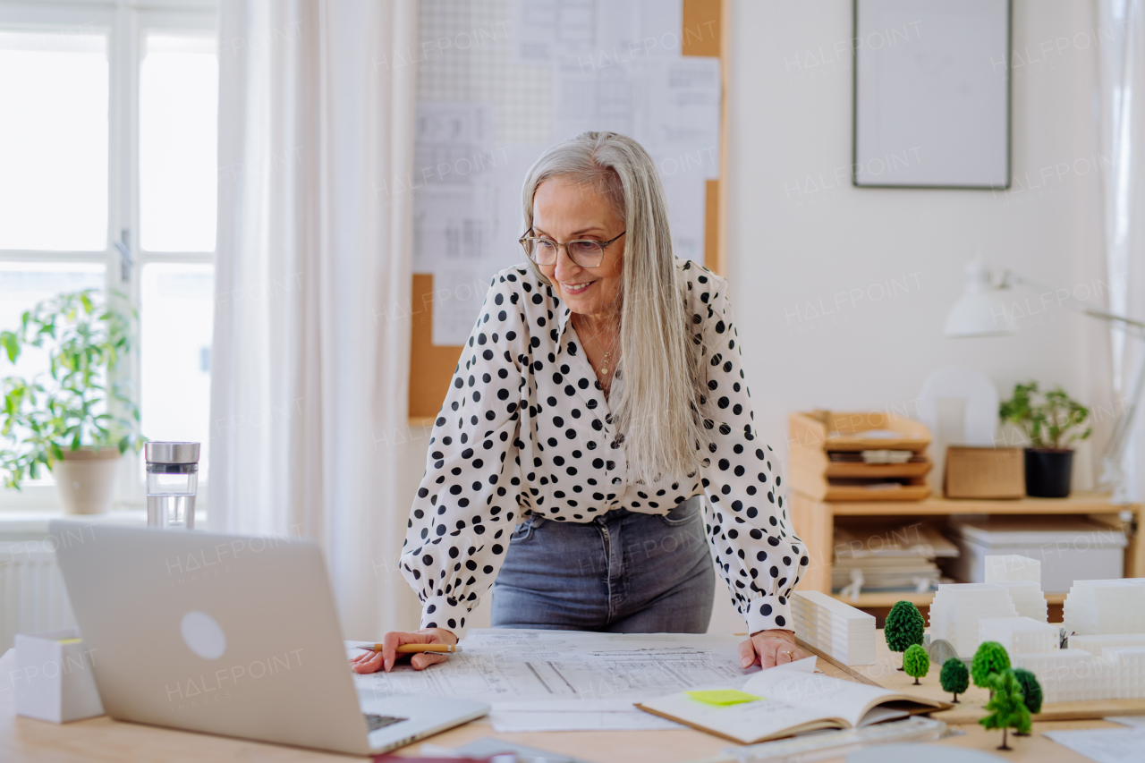 A senior woman architect with model of houses looking at laptop in office.