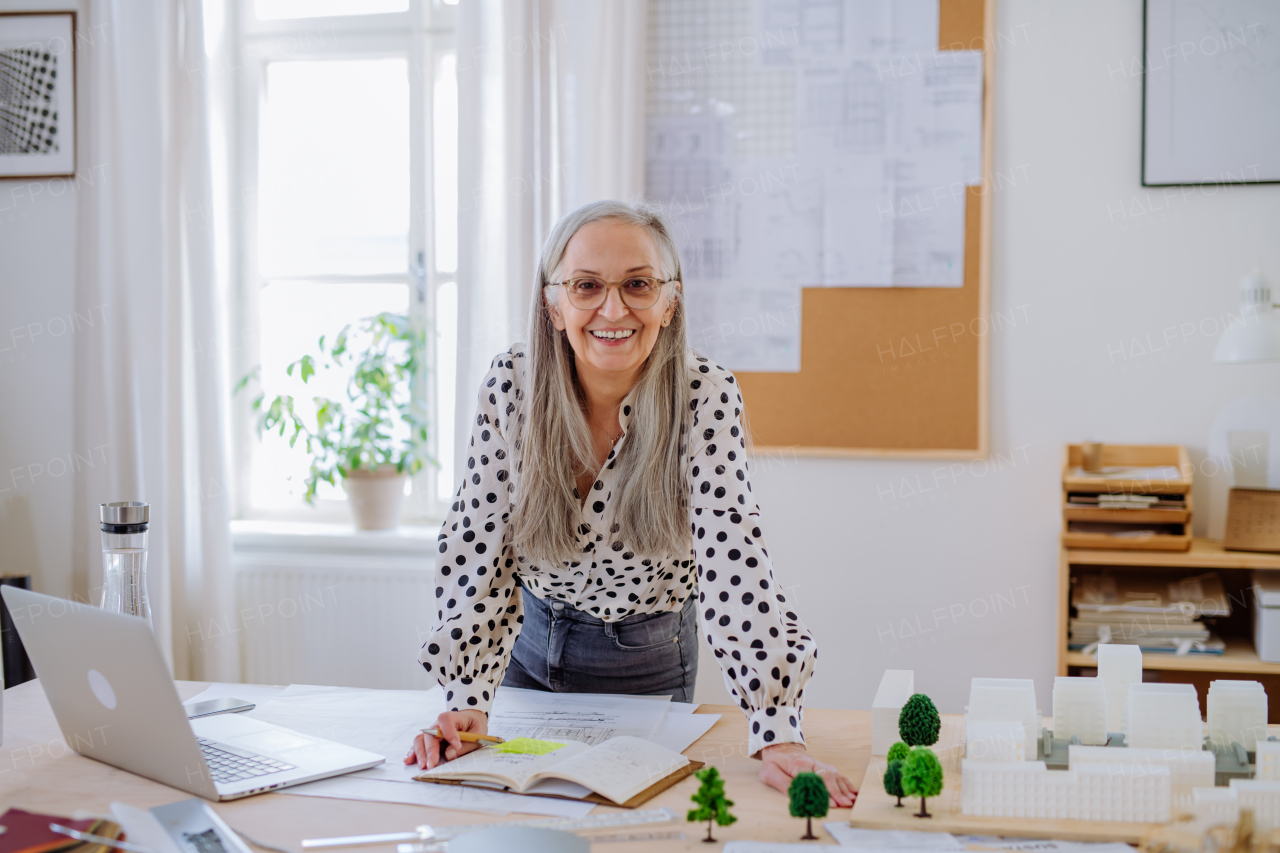 Happy senior woman architect with model of houses standing in office and looking at camera.