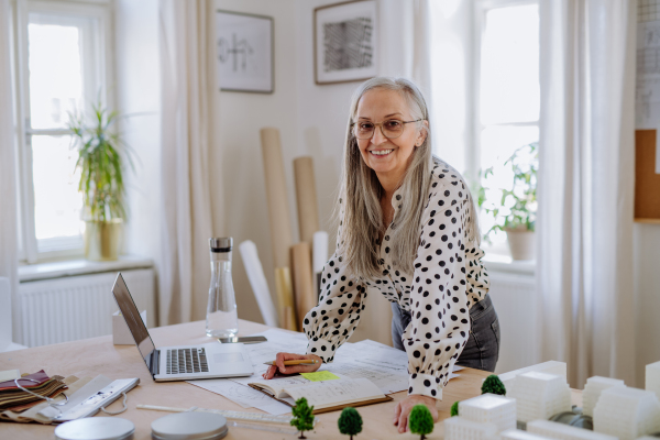 Happy senior woman architect with model of houses standing in office and looking at camera.