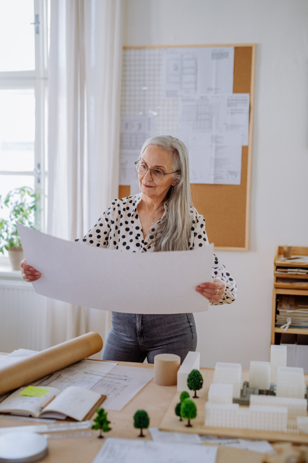 A senior woman architect with model of houses looking at blueprints in office.