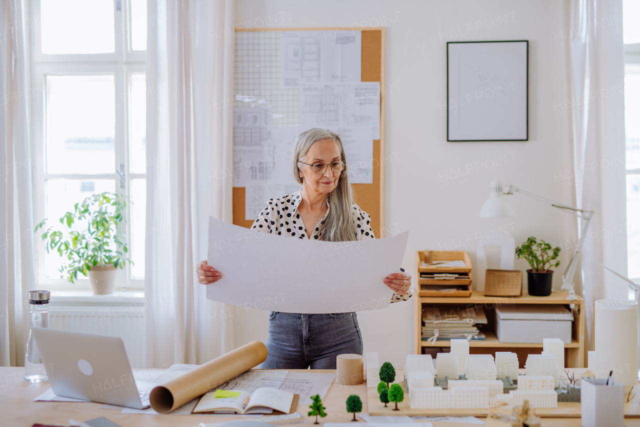 A senior woman architect with model of houses looking at blueprints in office.