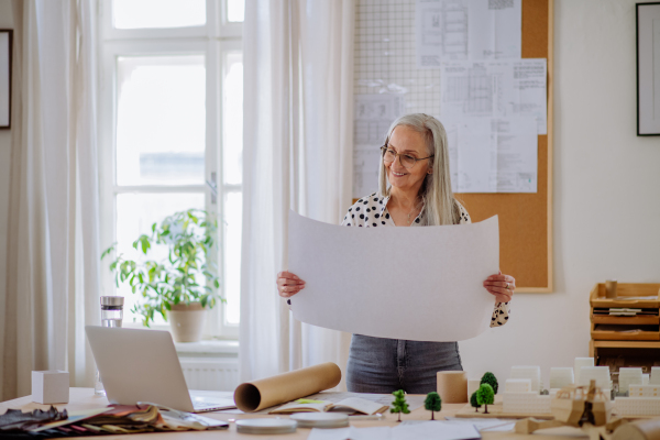 A senior woman architect with model of houses looking at blueprints in office.