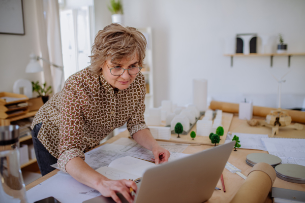 A senior woman architect with model of houses looking at blueprints in office.