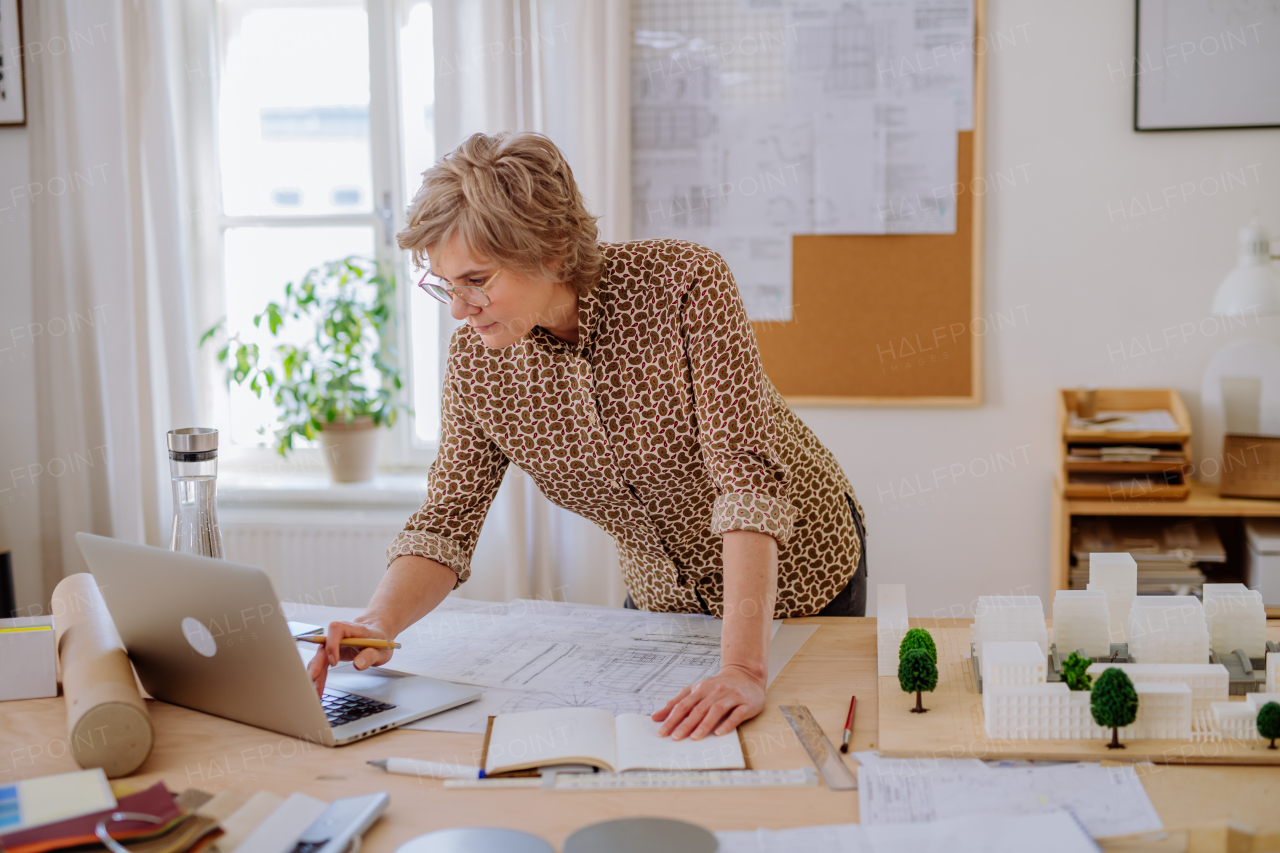 A senior woman architect with model of houses looking at blueprints in office.