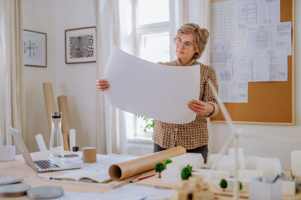 A senior woman architect with model of houses looking at blueprints in office.