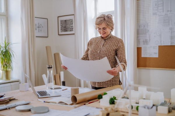 A senior woman architect with model of houses looking at blueprints in office.