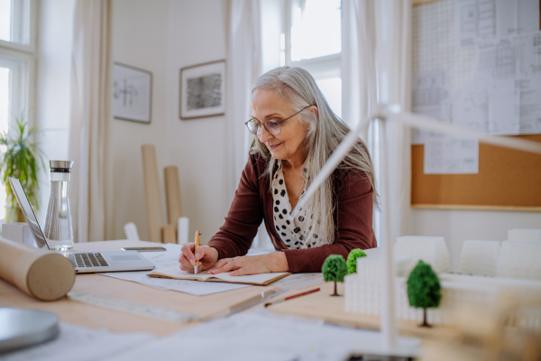 A senior woman architect with model of houses standing in office and and writing notes.