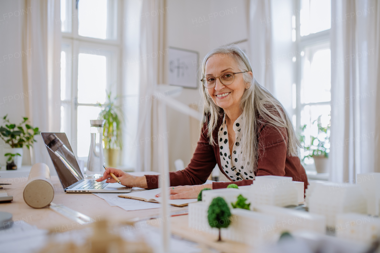 Happy senior woman architect with model of houses working on laptop in office and looking at camera.