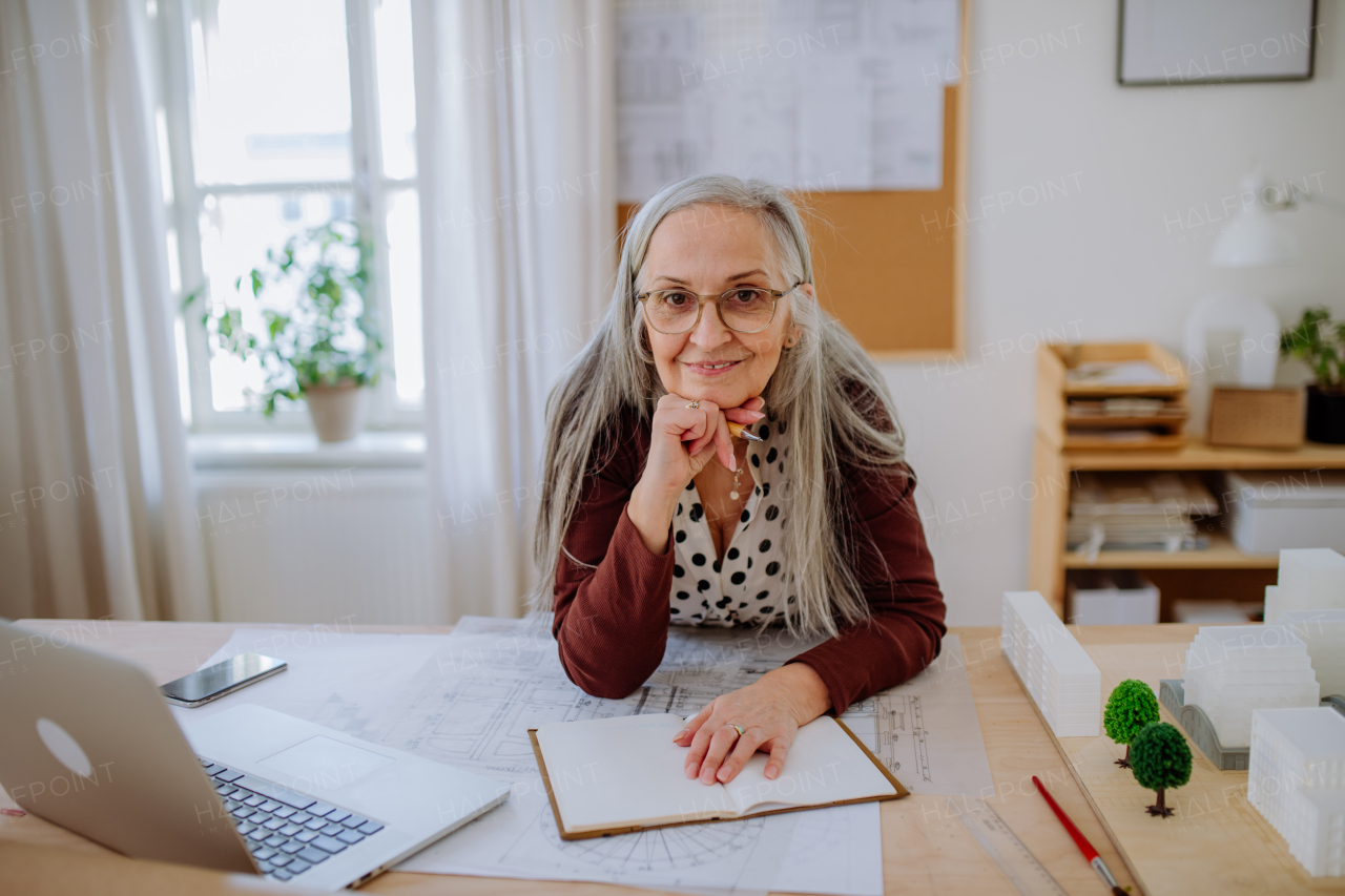 Happy senior woman architect with model of houses standing in office and looking at camera.