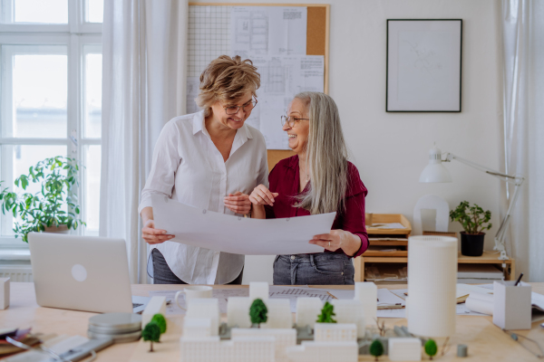 Mature women eco architects with model of modern bulidings and blueprints working together in an office.