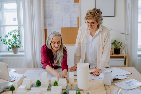 Mature women eco architects with model of modern bulidings and blueprints working together in an office.