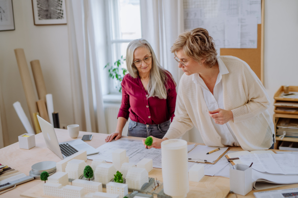 Mature women eco architects with model of modern bulidings and blueprints working together in an office.