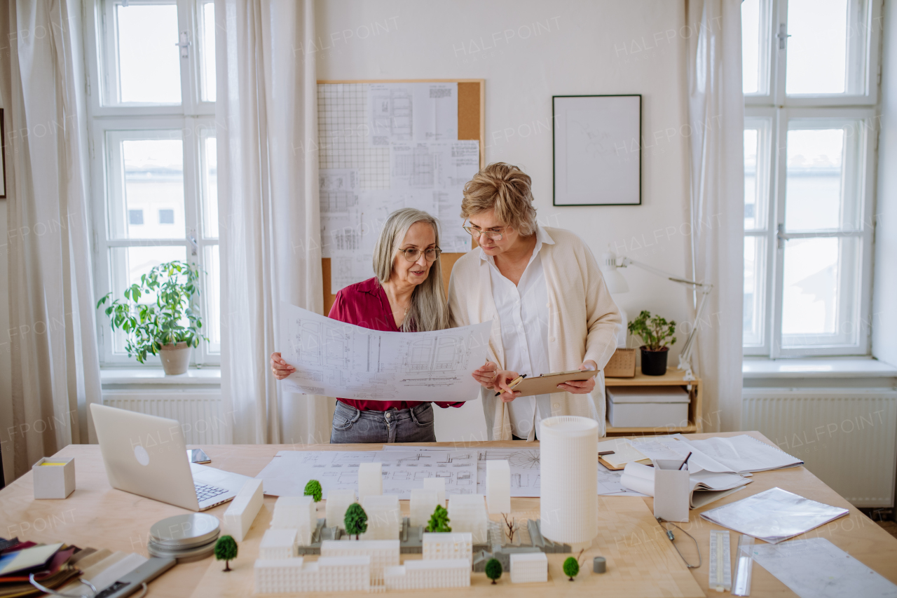 Mature women eco architects with model of modern bulidings and blueprints working together in an office.