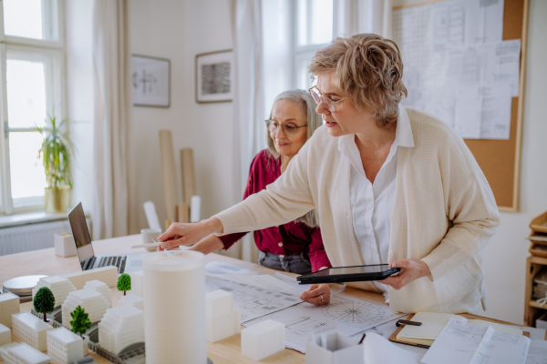 Mature women eco architects with model of modern bulidings and blueprints working together in an office.