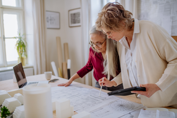 Mature women eco architects with model of modern bulidings and blueprints working together in an office.