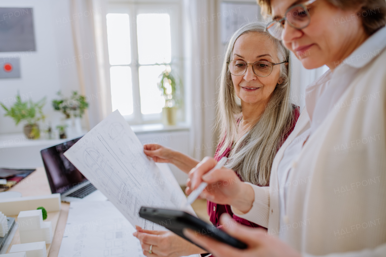 Mature women eco architects with blueprints working on a tablet together in office.