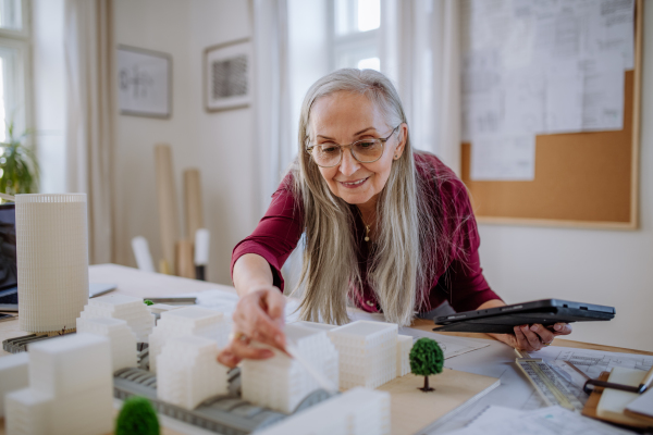 A senior woman eco architect with model of modern bulidings working in office.