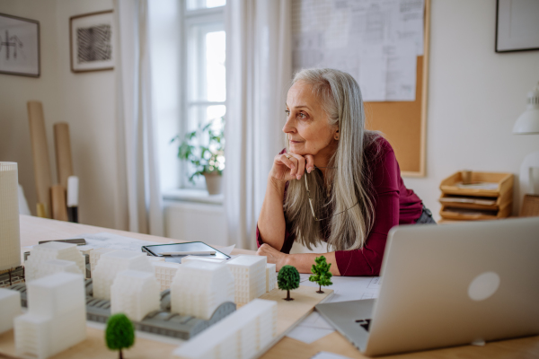 A senior woman eco architect with model of modern bulidings working in office.